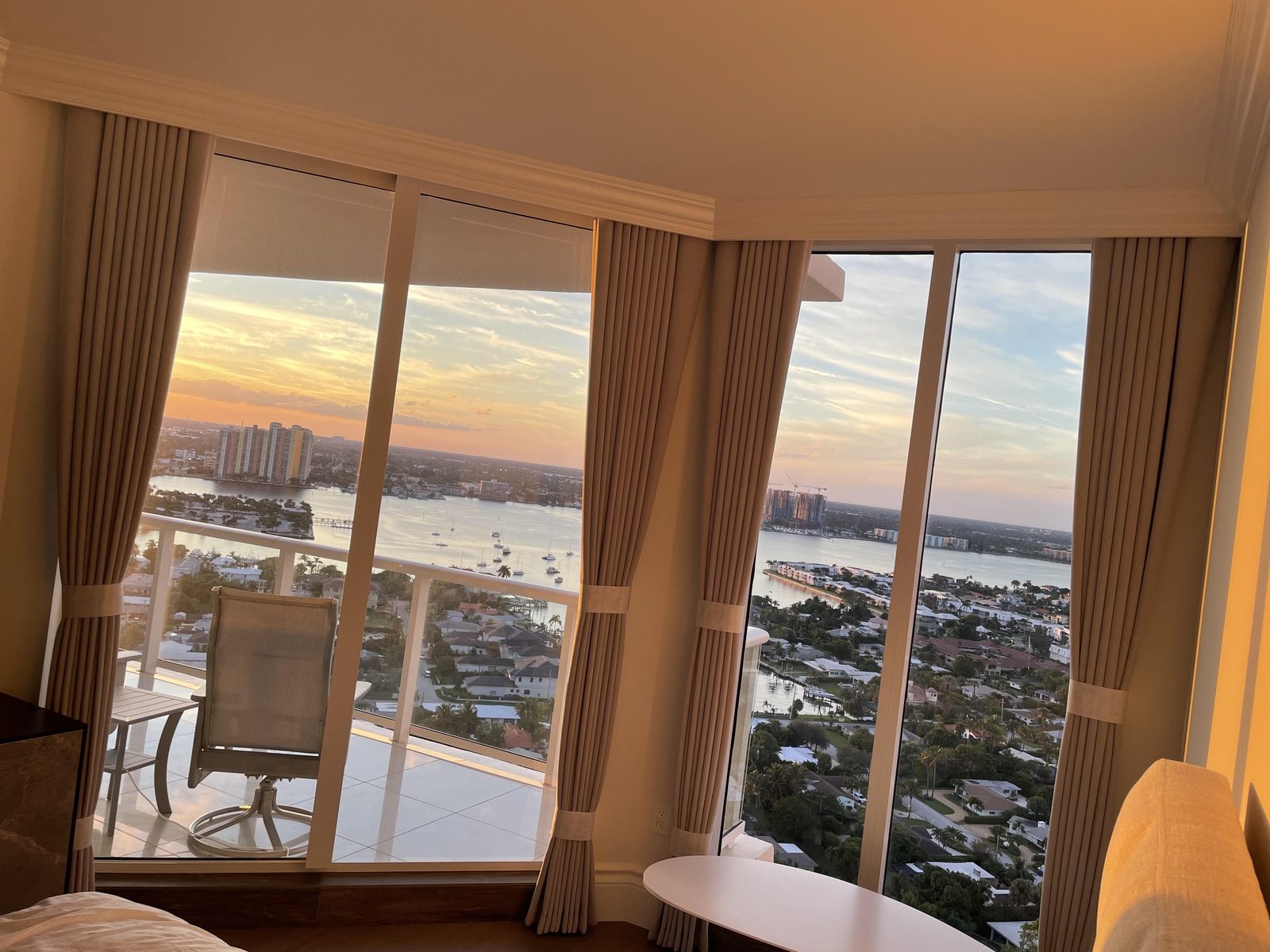 Condo bedroom corner windows at sunset with beige drapery panels and roller shades